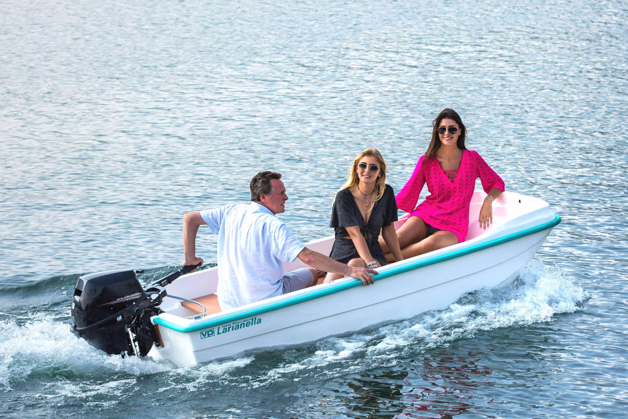 Tre persone che navigano sulla Larianella. La barca è in movimento su un'acqua calma e limpida. Una delle persone, un uomo, è seduto nella parte posteriore della barca e manovra il motore. Le altre due persone, entrambe donne, sono sedute al centro e nella parte anteriore della barca, una con un vestito nero e l'altra con un vestito rosa. Tutti sembrano rilassati e godono della gita in barca.