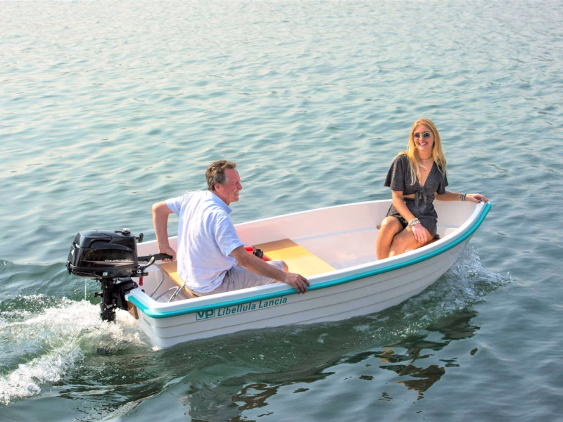 Due persone che navigano su una Libellula bianca a motore. La barca è in movimento su un'acqua calma e limpida. Un uomo è seduto nella parte posteriore della barca e manovra il motore, mentre una donna è seduta al centro della barca, indossando un vestito nero. Entrambi sembrano rilassati e godono della gita in barca, con il sole che illumina la scena e crea un'atmosfera serena.