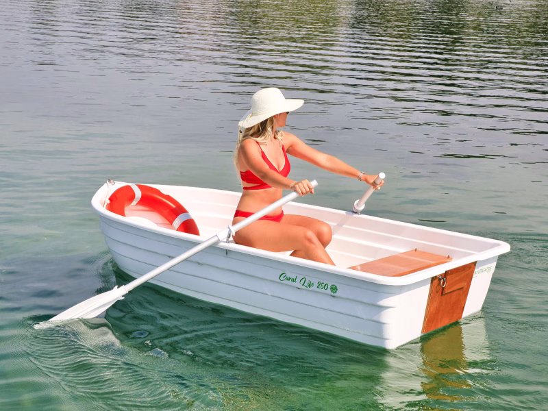 Una barca bianca di tipo tender, modello Coral Life 250, realizzata in plastica riciclata e completamente riciclabile. Una donna in costume da bagno rosso e cappello bianco è seduta all'interno della barca, remando con remi bianchi. All'interno della barca c'è anche un salvagente rosso e bianco. La barca ha sedili in legno e un design semplice e funzionale. L'ambiente circostante è sereno, con acque calme e un'atmosfera rilassata.
