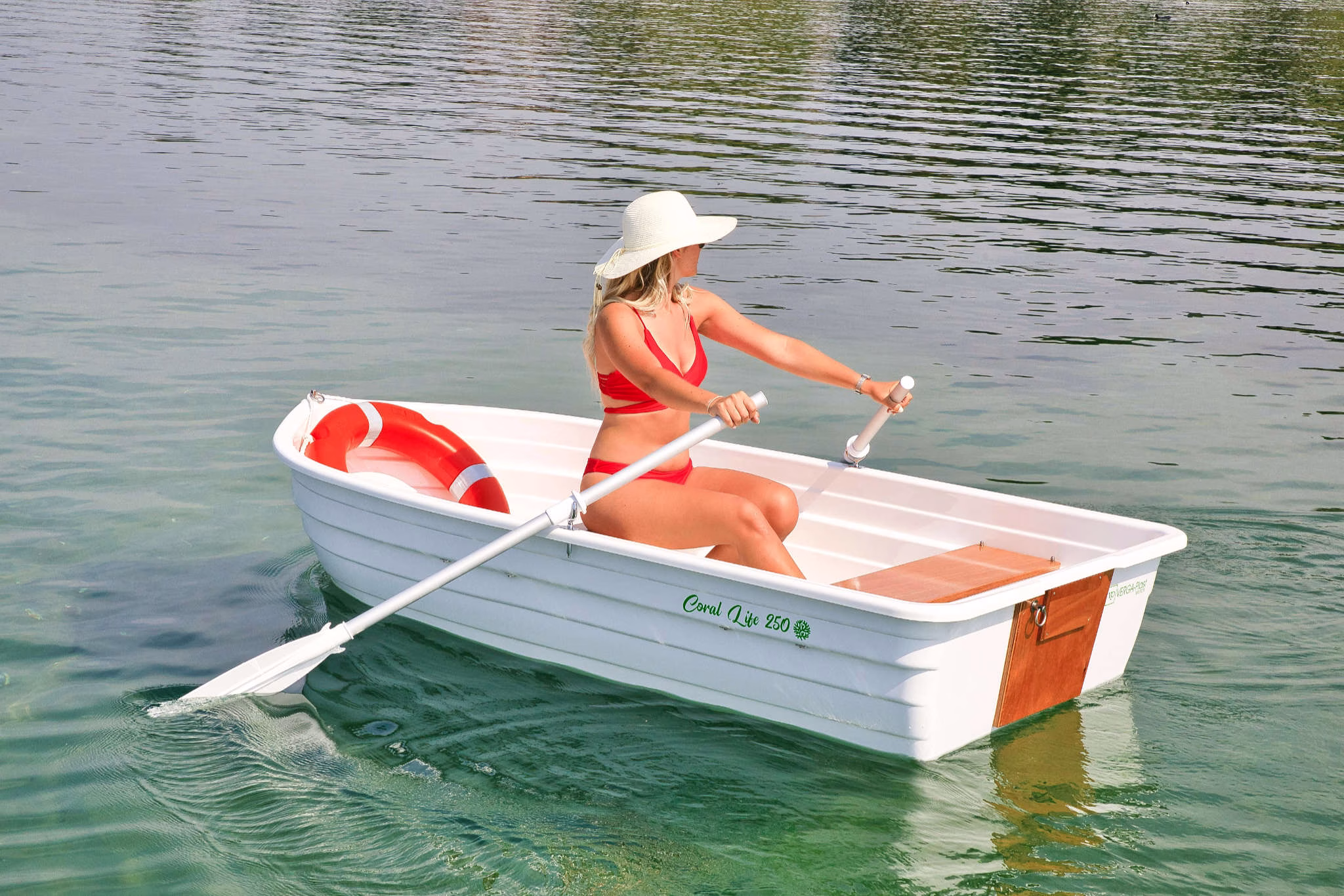 Una barca bianca di tipo tender, modello Coral Life 250, realizzata in plastica riciclata e completamente riciclabile. Una donna in costume da bagno rosso e cappello bianco è seduta all'interno della barca, remando con remi bianchi. All'interno della barca c'è anche un salvagente rosso e bianco. La barca ha sedili in legno e un design semplice e funzionale. L'ambiente circostante è sereno, con acque calme e un'atmosfera rilassata.