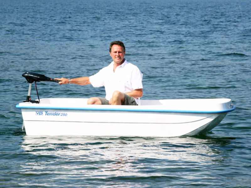 Un uomo che naviga da solo su un Tender 250 bianco a motore in un'acqua calma e limpida. L'uomo è seduto nella parte posteriore della barca, indossa una maglietta bianca e sorride verso la fotocamera mentre manovra il motore. Il cielo è sereno, creando un'atmosfera tranquilla e rilassata durante la gita in barca.