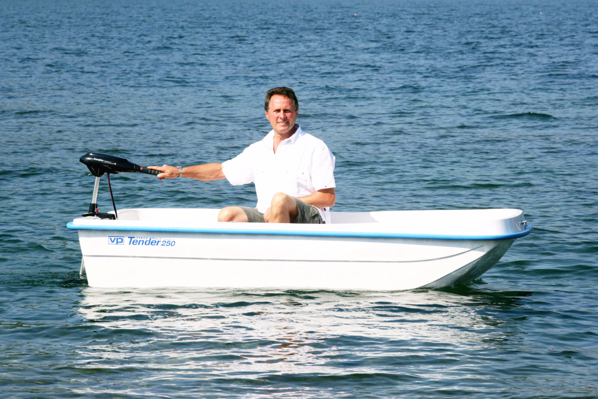 Un uomo che naviga da solo su un Tender 250 bianco a motore in un'acqua calma e limpida. L'uomo è seduto nella parte posteriore della barca, indossa una maglietta bianca e sorride verso la fotocamera mentre manovra il motore. Il cielo è sereno, creando un'atmosfera tranquilla e rilassata durante la gita in barca.